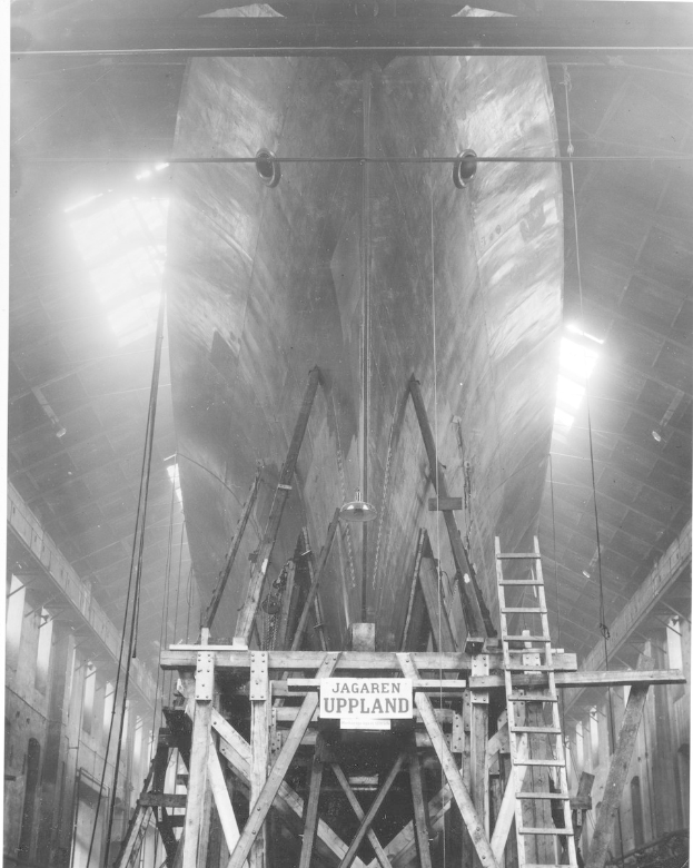 Großes Schwarz-Weiß-Foto eines Schiffs in einem Schiffbauhafen, das eine Leiter, hölzerne Pfosten, ein Schild, Metallrahmen, Deckenleuchten und Fenster zeigt.