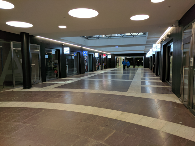 Ein langer, gut beleuchteter Flur mit Menschen, umgeben von Säulen und Glastüren, und mit Schautafeln mit Text, ähnlich einem belebten Rathauses.