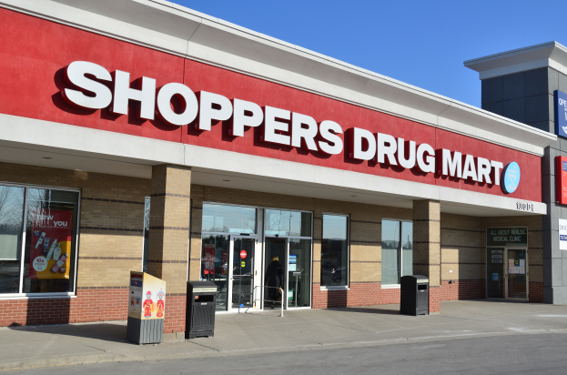Ein Schaufenster einer Drogerie mit der Aufschrift "Shoppers Drug Mart" und einer Straße davor, mit zwei Mülltonnen im Vordergrund und einem klaren blauen Himmel im Hintergrund.
