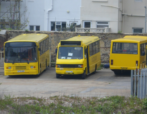 Drei gelbe Busse nebeneinander auf einem eingezäunten Parkplatz mit Grünfläche, einem Gebäude und Rohren im Hintergrund.