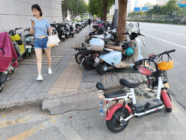 Eine Frau in einem blauen T-Shirt, blauen Shorts und weißen Schuhen geht auf einem Bürgersteig, hält ein Handy in einer Hand und eine Tasche in der anderen, vorbei an einer Reihe geparkter E-Scooter mit Bäumen, Gebäuden und einem klaren blauen Himmel im Hintergrund.