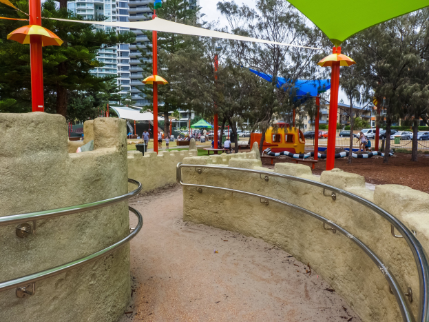 Spielplatz mit einem grünen und gelben Dach, umgeben von Zäunen, Pfählen, Zelten, Bäumen, Fahrzeugen, Menschen und Gebäuden unter einem klaren blauen Himmel.