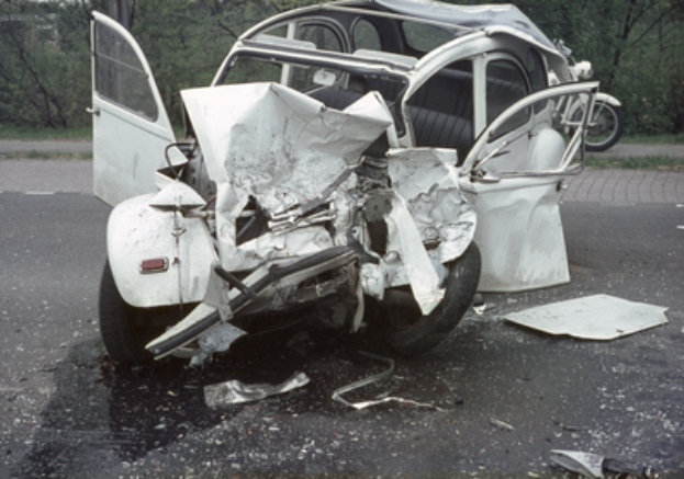 Ein stark beschädigtes Auto mit schwerer Frontpartie steht am Straßenrand, im Hintergrund sind Bäume, ein Gebäude und ein weiteres Fahrzeug zu sehen.