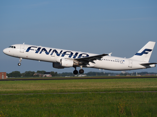 Ein Airbus A320-200 der Finnair startet vom Frankfurter Flughafen, mit Gras darunter, Gebäuden und Bäumen im Hintergrund und einem klaren blauen Himmel darüber.
