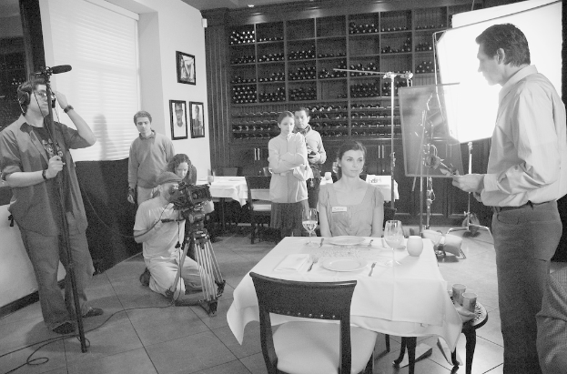 Ein Filmset mit einem Mann auf der rechten Seite, einer Frau an einem Tisch mit Tellern, Gabeln und Gläsern in der Mitte und zwei Personen, die eine Kamera bedienen, auf der linken Seite, mit Regalen, Rahmen und einem Bildschirm im Hintergrund.