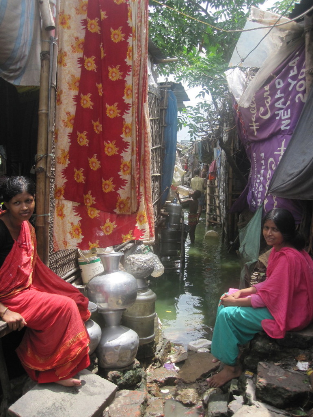 Zwei Frauen auf einer Bank in einem Slum-Gebiet sitzend, mit Stahlgegenständen und aufgehängter Wäsche in der Nähe und Häusern und Bäumen im Hintergrund.