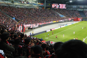 Ein Fussballspiel in einem grossen Stadion mit Spielern auf dem Feld und Zuschauern in den Rängen, umgeben von Bannern, einem Zaun, einem Display, Metallstangen und einem Dach mit Deckenlampen.