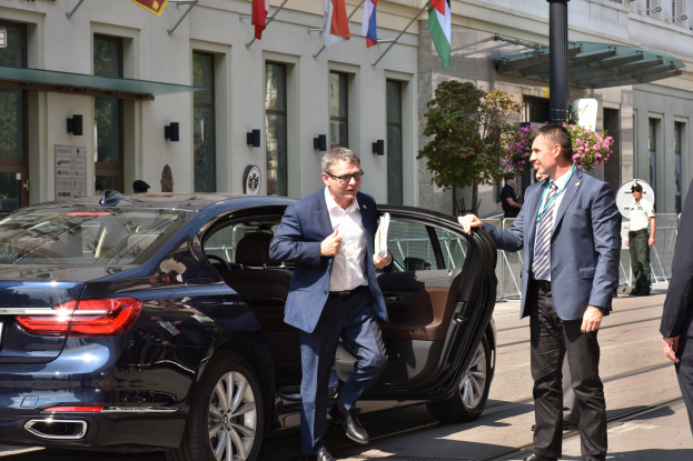 Eine Gruppe von Männern in Anzügen und Krawatten steht neben einem schwarzen Auto auf einer Straße, umgeben von Gebäuden, Fahnen, Pfählen, Bäumen, Pflanzen und Blumen, scheinbar über die neue BMW 7er-Reihe diskutierend.