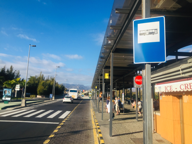 Busbahnhof mit einem Unterstand und einem Schild, Fahrzeuge und Fußgänger auf der Straße, Bäume, Laternenpfähle, Berge und einen klaren blauen Himmel im Hintergrund.