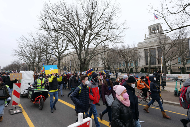 Eine große Gruppe von Menschen marschiert in einem Protest durch eine Straße in Washington, D.C., mit Schildern, Bannern und Fahrrädern, mit Bäumen, Schildern und einem klaren blauen Himmel im Hintergrund.