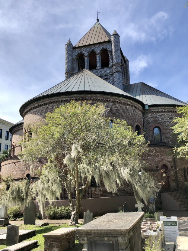 Außenansicht der Kirche des Heiligen Grabes in Charleston, South Carolina, mit einem Turm, einem Baum, umgeben von Gebäuden mit Fenstern, einer Treppe mit Geländer und einem wolkenverhangenen Himmel über Gras und einem Schild unten.