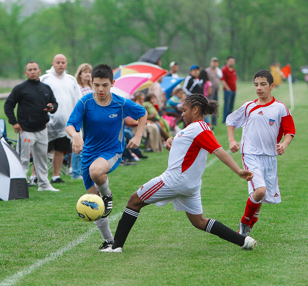 Drei Personen spielen Fußball mit einem gelben Ball, während eine Gruppe von Menschen hinter ihnen steht und sitzt und Schirme hält, mit Bäumen im Hintergrund.