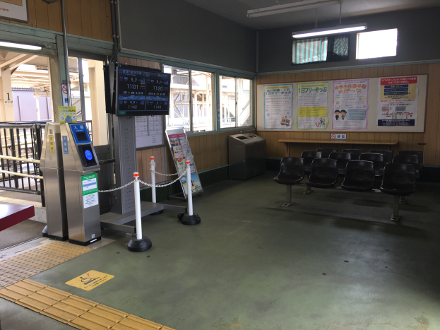 Ein belebter Bahnhofseingang mit Sitzgelegenheiten, einer Informations counter, Ticketautomaten, Kettenbarrieren, Informationsanzeigen, Wandplakate, Deckenleuchten und Geländern.