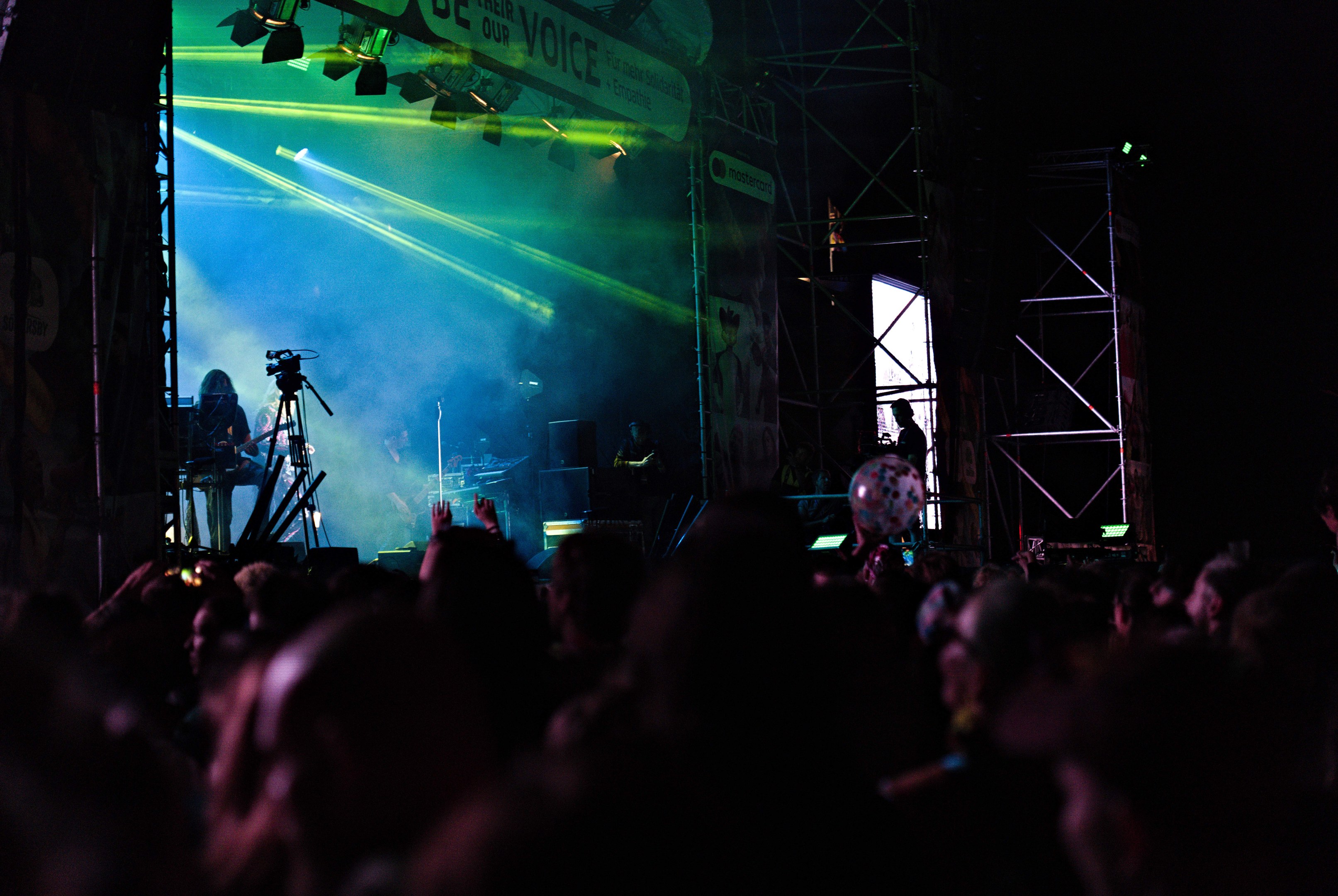 Eine Menge schaut einem Live-Konzert auf einer hell erleuchteten Bühne mit Musikern, Mikrofonen und einem Banner im Hintergrund unter dunklen Umständen zu.