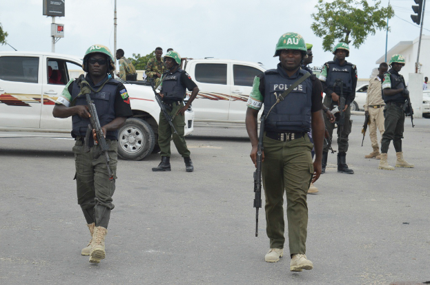 Eine Gruppe von Polizisten in Helmen, die Waffen halten, geht eine Straße entlang mit Fahrzeugen, Laternen, Ampeln, Texttafeln, Bäumen, Gebäuden und einem bewölkten Himmel im Hintergrund.