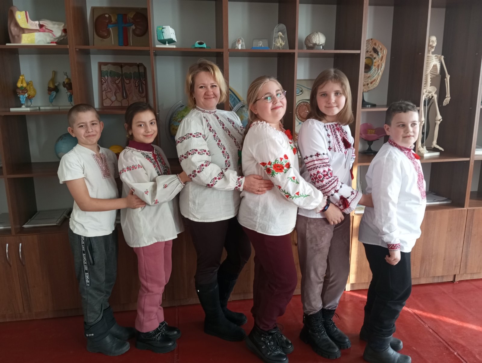 Eine Gruppe von Kindern in traditioneller ukrainischer Kleidung vor einem Regal mit Spielsachen, einem Skelett und anderen Gegenständen, das wohl eine ukrainische Schule ist.
