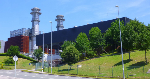 Ein großes Gebäude mit zahlreichen Fenstern, als Kraftwerk durch Beschilderung identifiziert, umgeben von Straßeninfrastruktur, Fahrzeugen, Grünflächen und einem bewölkten Himmel.