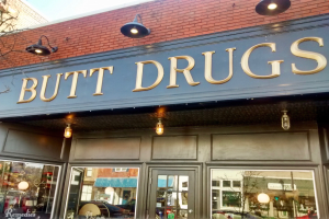 Außensicht eines Gebäudes mit einer beleuchteten Schrift 'butt drugs' und Glaswänden, die umliegende Fahrzeuge, Gebäude, Bäume und Himmel zeigen, was auf einen neu öffneten Drogengeschäft hindeutet.