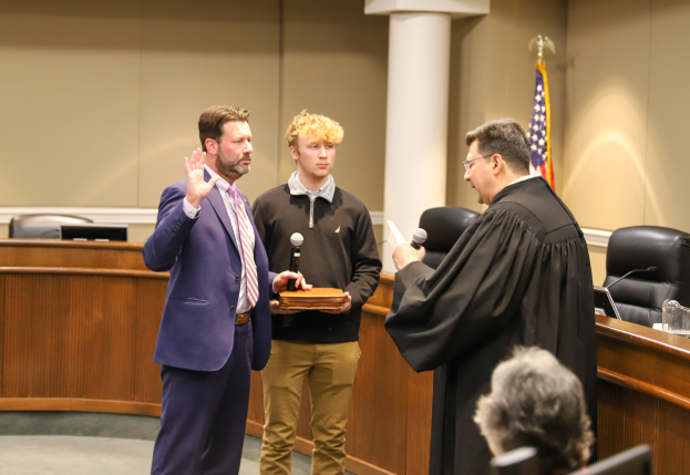 Ein Mann im Anzug und mit Krawatte steht neben einem Richter in einer schwarzen Robe in einem Gerichtssaal, hält ein Mikrofon und ein Buch in der Hand, mit Stühlen, Tischen, einer Flagge, einer Säule und einer Wand im Hintergrund und einer Person, die in einem Stuhl am unteren Bildrand sitzt.