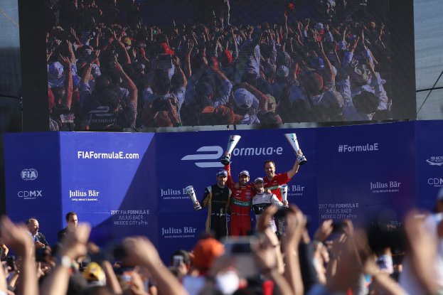 Gruppe von Menschen auf einem Formel-E-Podium in Mexiko-Stadt mit Pokale, Sponsor-Wänden und einem Hintergrundbild.