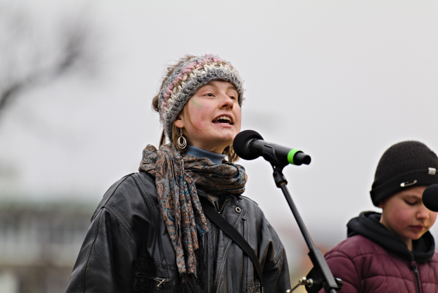 Eine Frau in einer schwarzen Jacke, einem Schal und einer Mütze singt in ein Mikrofon auf der Bühne, neben einer Person in einer maronenfarbenen Jacke und einer schwarzen Mütze, während eine unscharfe Menge zusieht.