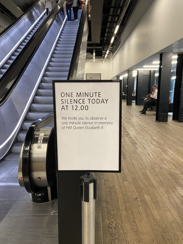 Eine Rolltreppe in einem Flughafen mit einem Schild, auf dem "Eine Minute Stille heute" steht, sowie ein paar Menschen darauf und Lampen an der Decke im Hintergrund.