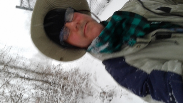 Eine Person in Jacke und Hut steht vor einem verschneiten Hintergrund mit Bäumen links unten.