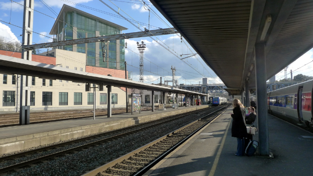 Eine Szene an einem Bahnhof mit Menschen, die auf der rechten Seite stehen und sitzen, einem Zug, der in der Mitte auf den Schienen fährt, und Gebäuden, Pfählen und einem bewölkten Himmel im Hintergrund.