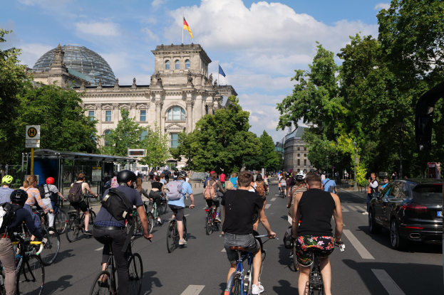 Eine Gruppe von Menschen, die auf Fahrrädern eine von Bäumen gesäumte Straße in Berlin, Deutschland, entlangfährt, mit Gebäuden und einer Bushaltestelle im Hintergrund, unter einem bewölkten Himmel und einer Flagge, die auf einem Gebäude weht.