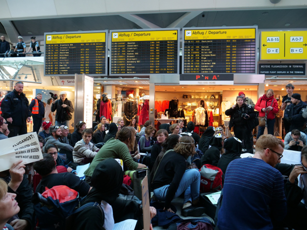 Große Menschenansammlung in einem Flughafen, einige sitzen mit Taschen und Papieren, andere stehen, mit Texttafeln und Schaufensterpuppen im Hintergrund, unter Deckenbeleuchtung, während einer Demonstration.