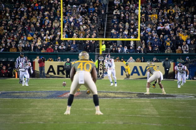 Fußballspieler in Sportkleidung und Helmen konkurrieren auf einem Feld mit Zuschauern im Hintergrund, Stadioninfrastruktur einschließlich Pfosten und Schilder mit Text sichtbar.