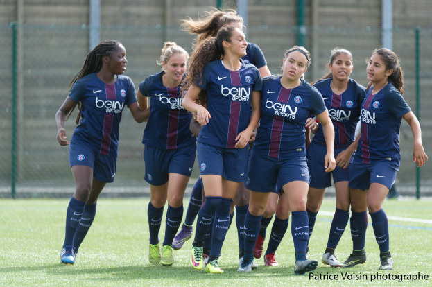 Eine Gruppe junger Frauen beim Fußballspielen auf einem Rasenplatz mit Maschendrahtzaun und Pfosten im Hintergrund und einer Wand dahinter, mit dem Text "Paris Saint-Germain Women's Soccer" in der rechten unteren Ecke.