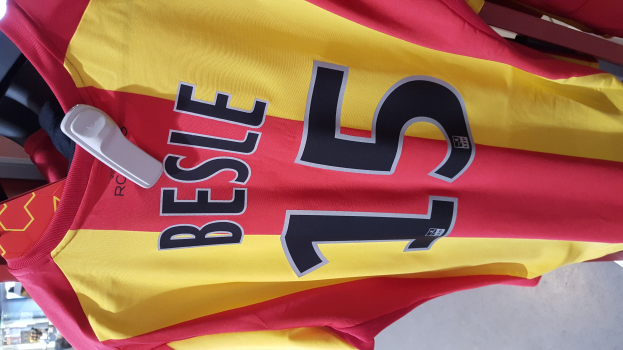 Ein rotes und gelbes Fußballtrikot mit aufgedrucktem Text und Nummern, das an einer Ladengestell hängen, mit anderen Gegenständen und dem Boden im Hintergrund.