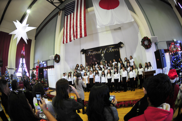 Eine Gruppe von Menschen auf einer Bühne mit festlichen Dekorationen, einem Podium und einer Menge darunter mit Kameras und Handys.