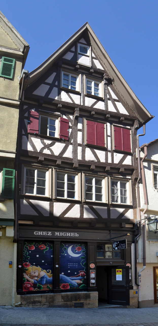 Altes Gebäude mit roten Fensterläden, mehreren Fenstern und klassischem Architekturstil, vor einem klaren blauen Himmel.