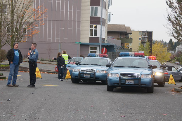 Autos auf einer Straße mit vier Menschen in der Nähe, Gebäude mit Fenstern im Hintergrund, Bäume und ein Notfall-Dreieck.