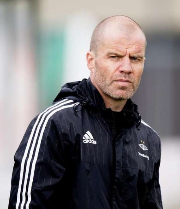 Kahlköpfiger Trainer in marineblauer Jacke mit weißen Streifen und weißem Kragen auf einem Fußballfeld stehend, Hände verschränkt und ernster Gesichtsausdruck.