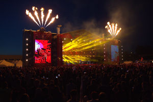 Eine große Menschenmenge versammelte sich auf einem Musikfestival, mit einer Bühne in der Mitte, auf der Musiker von hellen Scheinwerfern beleuchtet wurden und von Bannern umgeben waren, Zelte links daneben und ein dunkler Himmel mit Feuerwerk im Hintergrund.