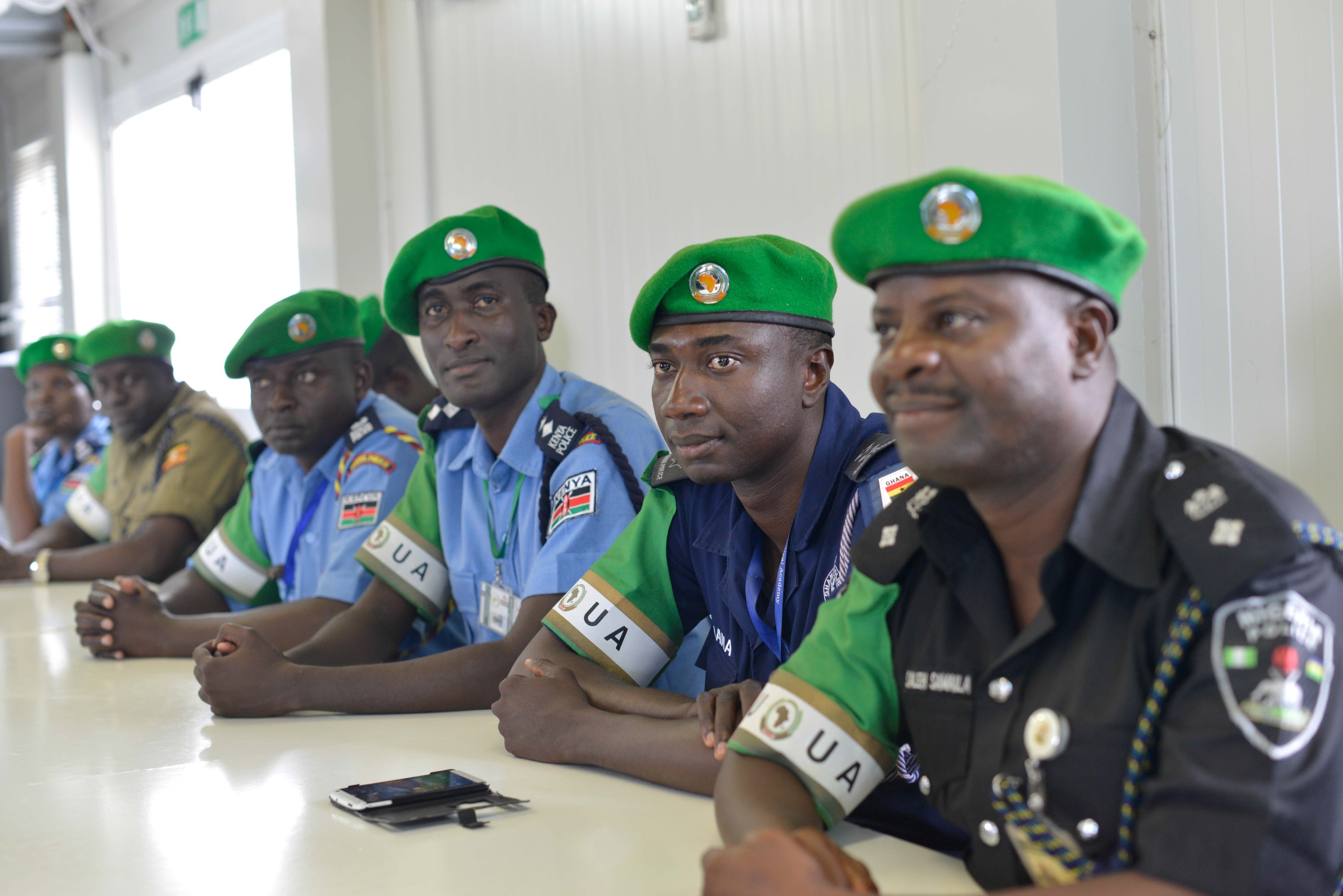 Gruppe von Polizeibeamten, die während einer Schulung an einem Tisch sitzen, mit Mobiltelefonen und anderen Gegenständen auf dem Tisch, in einem Raum mit Fenstern und Deckenbeleuchtung.