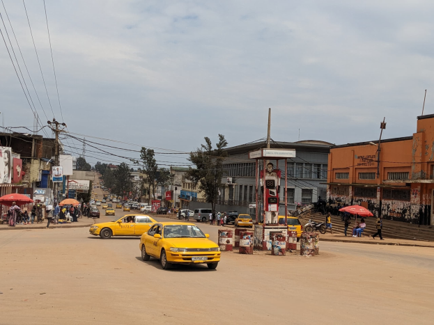 Ein gelbes Taxi fährt eine belebte Stadtstraße mit Gebäuden an den Seiten hinunter, während Fußgänger mit Regenschirmen auf dem Gehweg darunter unter bewölktem Himmel gehen.
