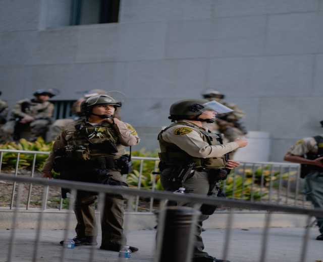 Eine Gruppe von Polizisten in Uniform, Helmen und Stiefeln steht vor einem Gebäude, einige halten Waffen, mit einem Metallzaun im Vordergrund und Pflanzen und einer Wand im Hintergrund.
