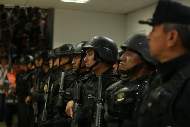 Gruppe von Polizisten in Uniform mit Helmen und Schusswaffen, die in Formation vor einer Wand mit Deckenbeleuchtung stehen.