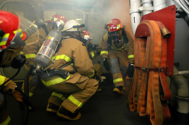 Feuerwehrleute in voller Schutzausrüstung mit Rauch aus dem Mund, stehend in einem raucherfüllten Raum mit Rohren und einer Wand im Hintergrund.