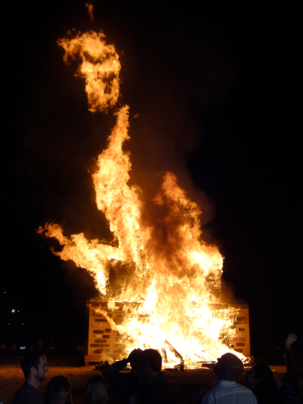 Ein Haus brennt mit ein paar Menschen in der Nähe, vor dunklem Hintergrund.