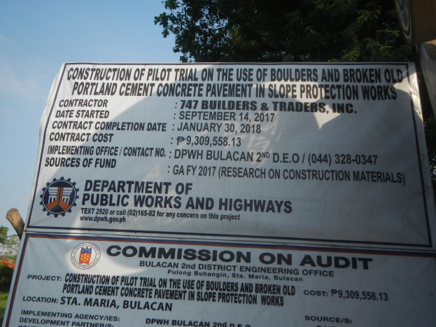 Schilder am Straßenrand mit der Aufschrift "Bauversuch zur Verwendung von Steinen und zerkleinertem altem Portlandzement-Beton in Hangschutzarbeiten" mit Bäumen, Pflanzen, Gras und einem klaren blauen Himmel im Hintergrund.