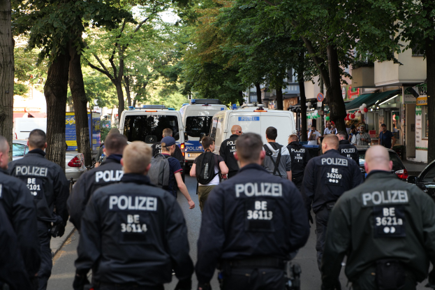 Polizeibeamte gehen eine von Bäumen gesäumte Straße mit geparkten Fahrzeugen und Passanten entlang.