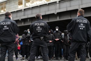 Eine Gruppe Von Polizisten In Uniform Steht Vor Einer Menge Von Menschen, Die Auch Schwarz Uniformen Und Masken Tragen, Mit Einer Brücke Und Einem Gebäude Im Hintergrund, Das Eine Stadtische Umgebung Vervielfältigt.