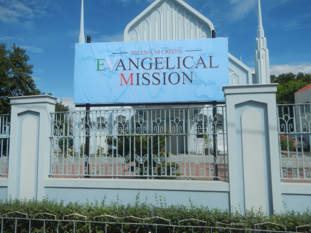 Eine Kirche mit einem Schild, auf dem "Evangelische Mission" steht, ist von einem Metallzaun, Pflanzen, Tieren und einem Haus mit einem Dach umgeben.