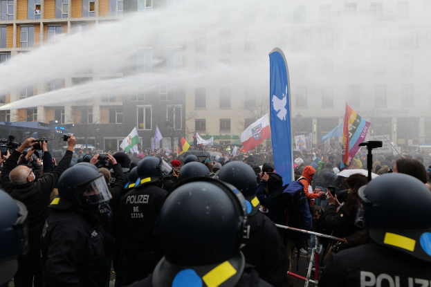 Eine Gruppe von Polizisten steht vor einer Menge, einige halten Kameras und tragen Helme, während ein Wasserwerfer Wasser auf sie sprüht, mit Gebäuden, Bäumen und einem klaren blauen Himmel im Hintergrund.