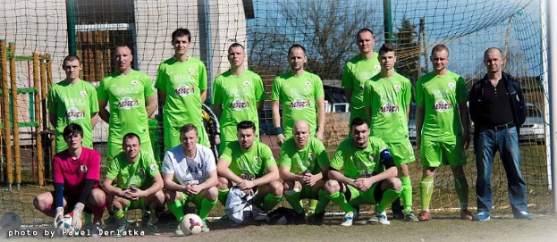Eine Gruppe von Männern in grünen T-Shirts steht auf einem Fußballfeld mit einem Ball vor ihnen, einige hocken sich, in der Nähe eines Torpfostens mit Bäumen, Gebäuden und Fahrzeugen im Hintergrund.